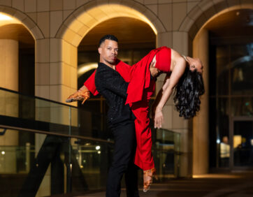 A close-up of salsa dancers at The Well — the woman, in a red dress, extends her leg over her partner’s shoulder while arching gracefully over the edge, supported confidently by the man in a black suit.