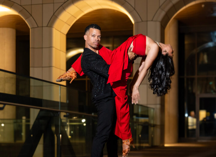 A close-up of salsa dancers at The Well — the woman, in a red dress, extends her leg over her partner’s shoulder while arching gracefully over the edge, supported confidently by the man in a black suit.
