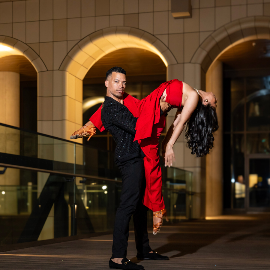 A close-up of salsa dancers at The Well — the woman, in a red dress, extends her leg over her partner’s shoulder while arching gracefully over the edge, supported confidently by the man in a black suit.