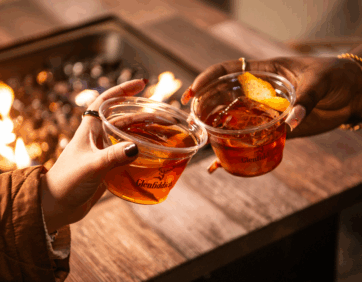 Two people clinking glasses of seasonal cocktails by a cozy fire at a holiday pop-up lounge in Toronto.